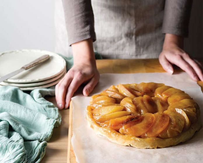 Origin of A Classic: Tarte Tatin on parchment with hands about to cut into tart